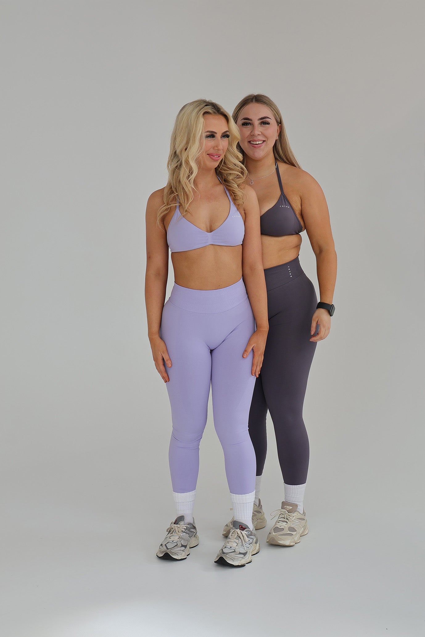 Two women smiling and standing side by side, wearing CNTOR seamless activewear sets in Storm and Lilac colors.