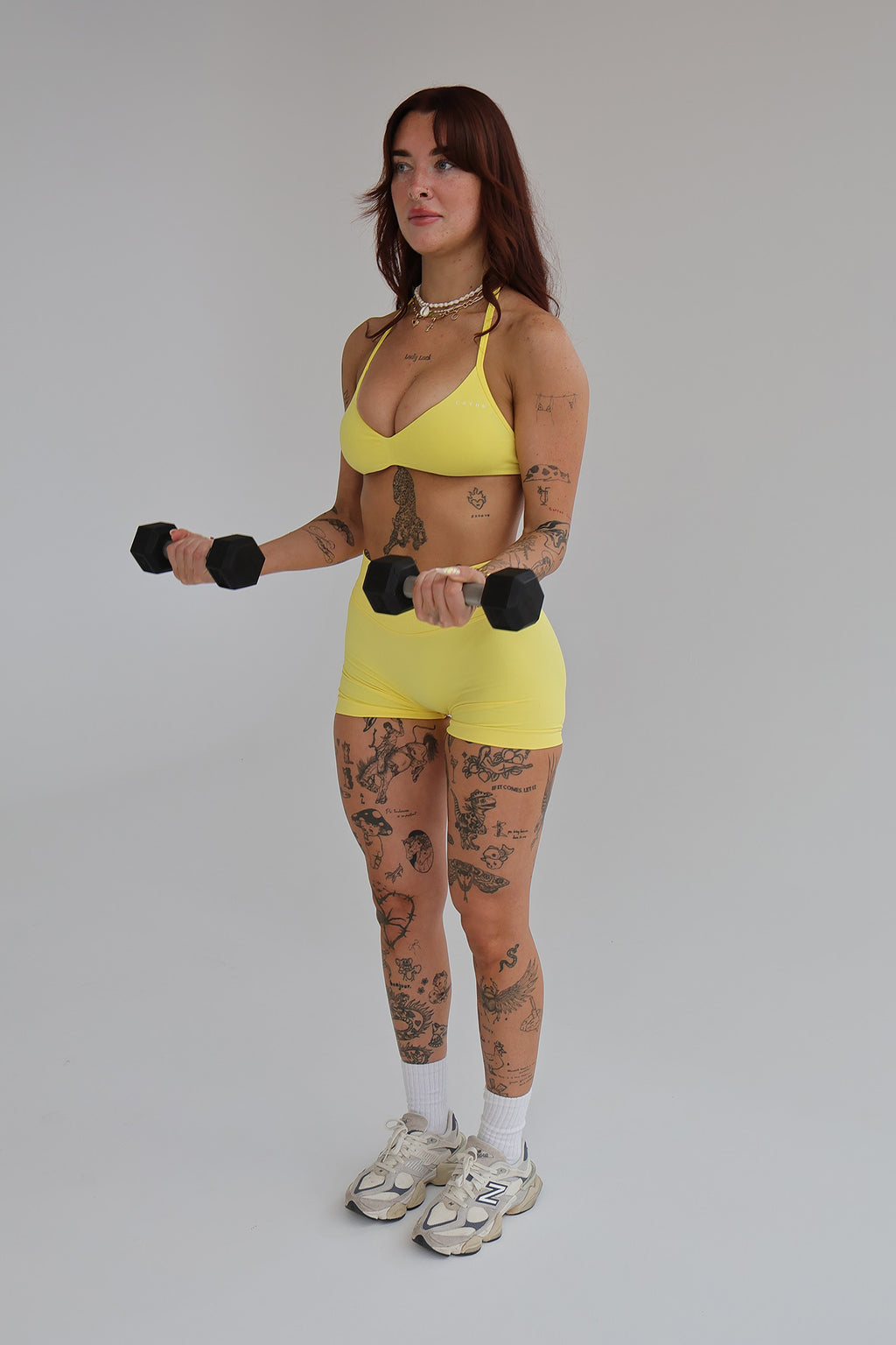 Woman wearing a cntor the label's yellow sports bra and shorts holding black dumbbells against a gray background