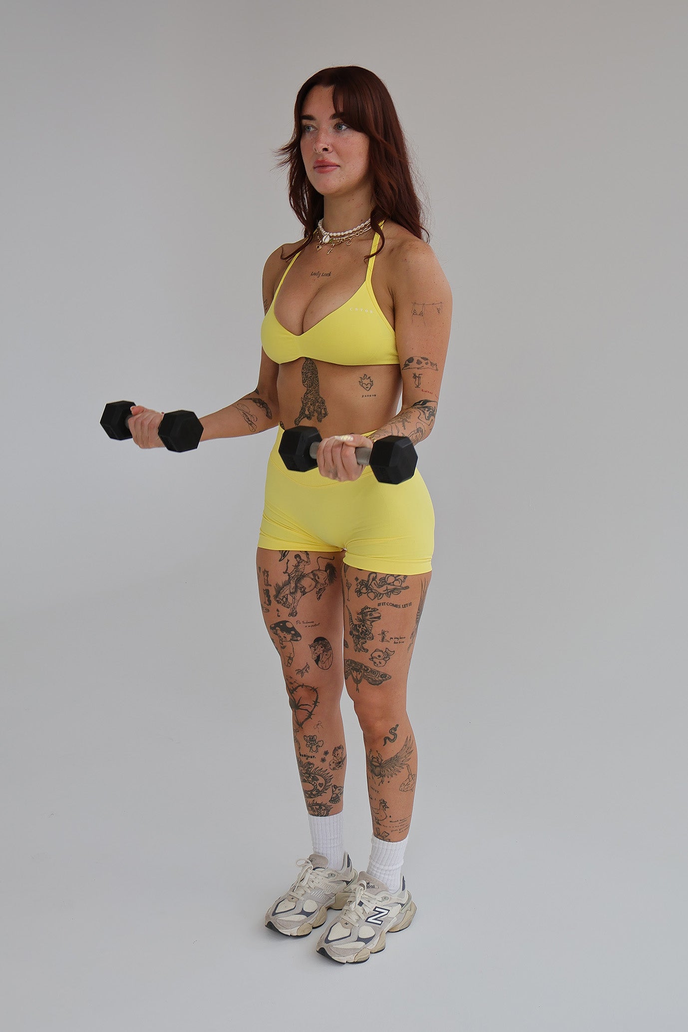 Woman wearing a cntor the label's yellow sports bra and shorts holding black dumbbells against a gray background