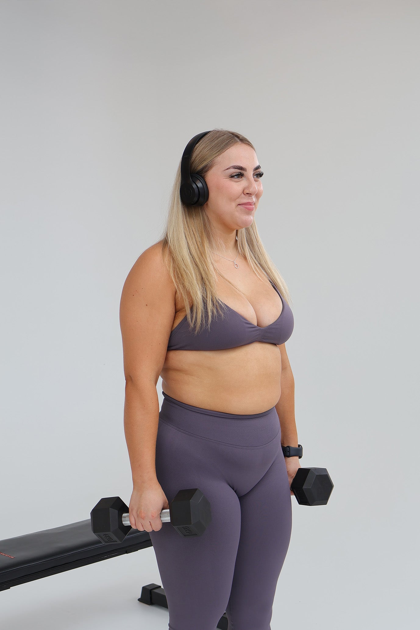 Woman in storm activewear attire with dumbbells and a bench on a plain background