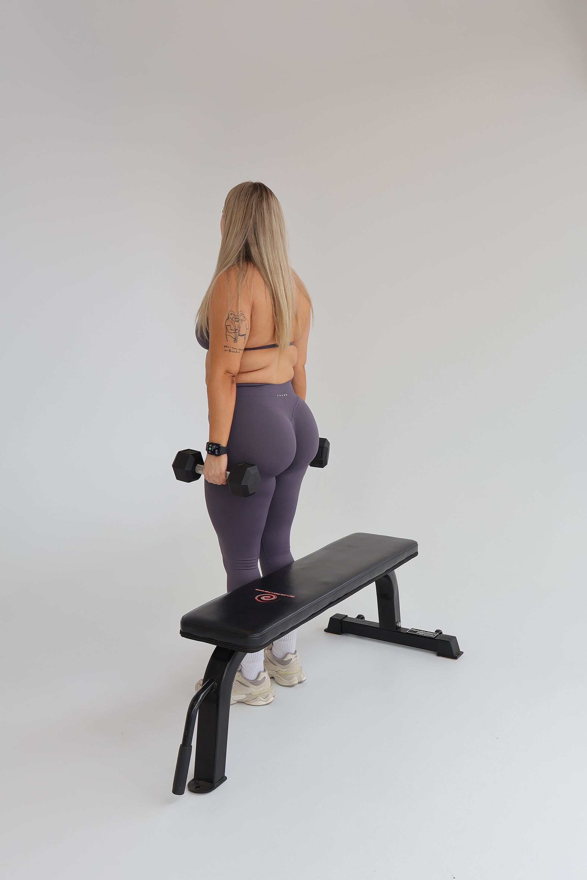 Woman wearing cntor the label activewear using a weight bench with dumbbells on a plain background