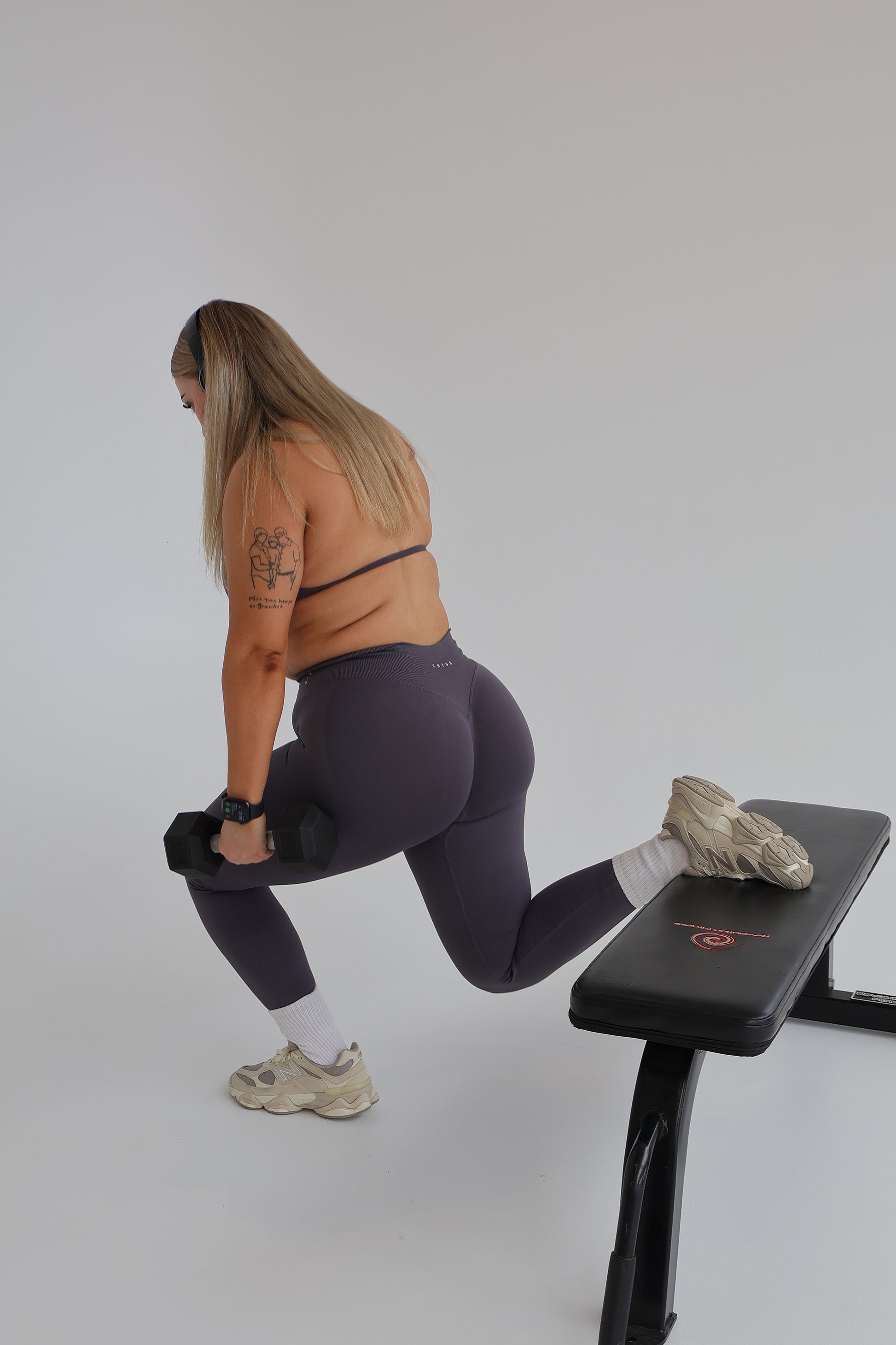 Woman wearing cntor the label activewear using a weight bench with dumbbells on a plain background
